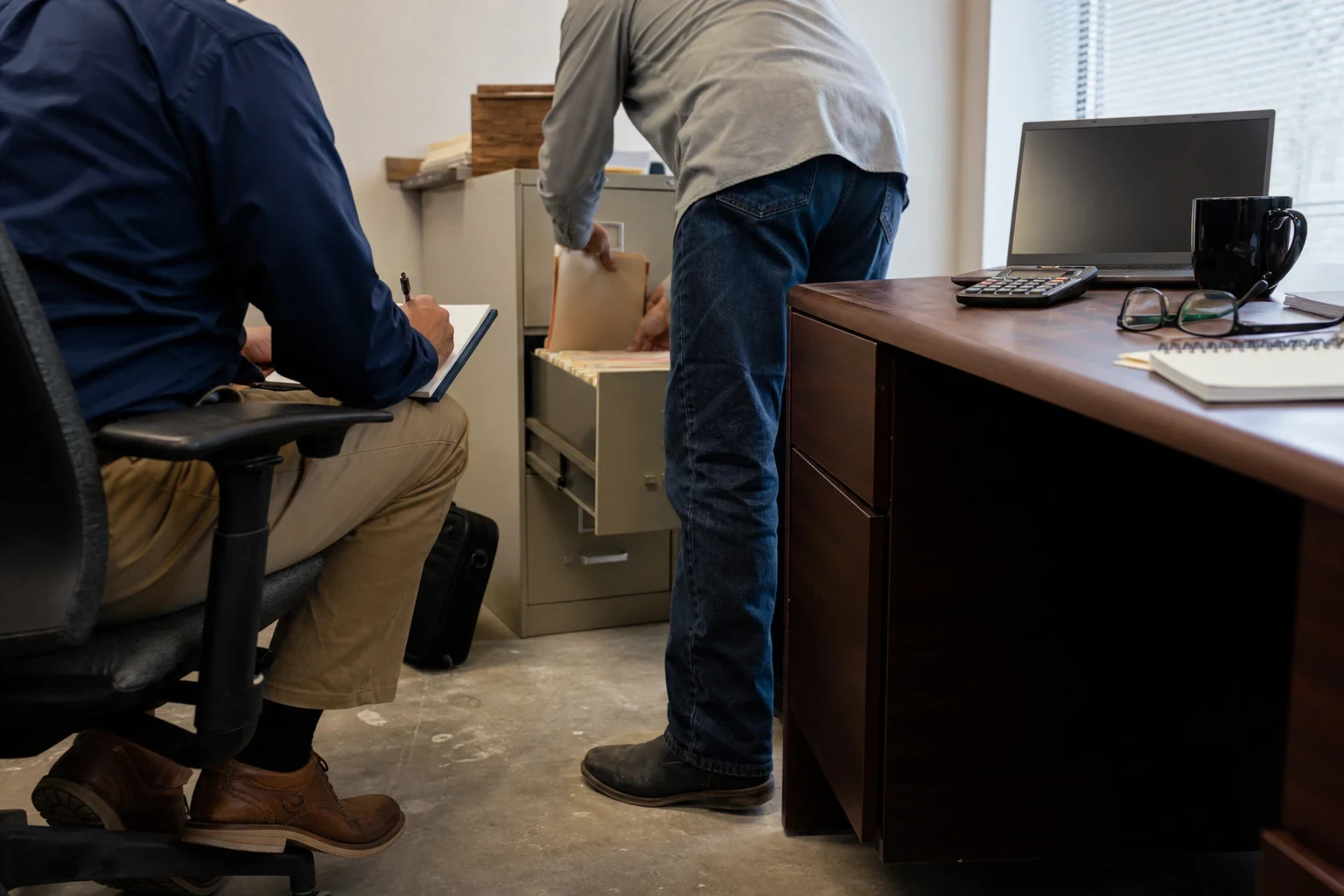 In a small office, a man pulls a file folder from a beige filing cabinet while a seated man takes notes beside a wooden desk holding a calculator, glasses and laptop.