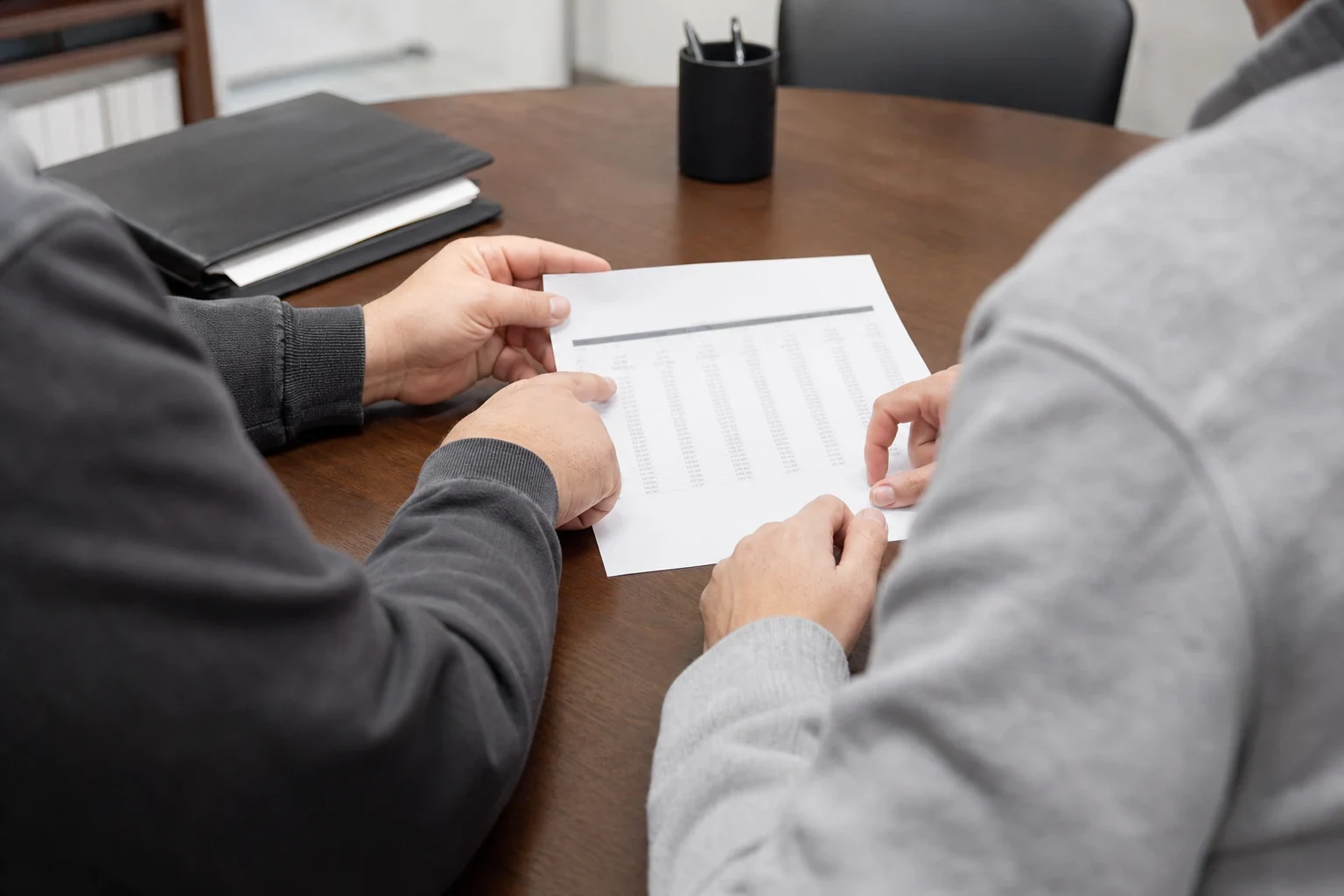 Two people at a round wooden conference table review a printed page of numeric figures, with a black binder and pen cup nearby.