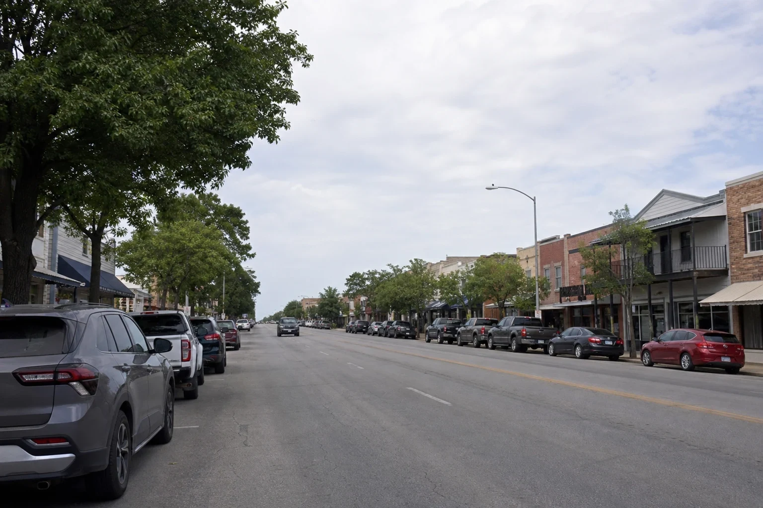 Wide main street lined with shade trees, brick storefronts with balconies and awnings, and pickups and cars parked along both curbs under an overcast sky